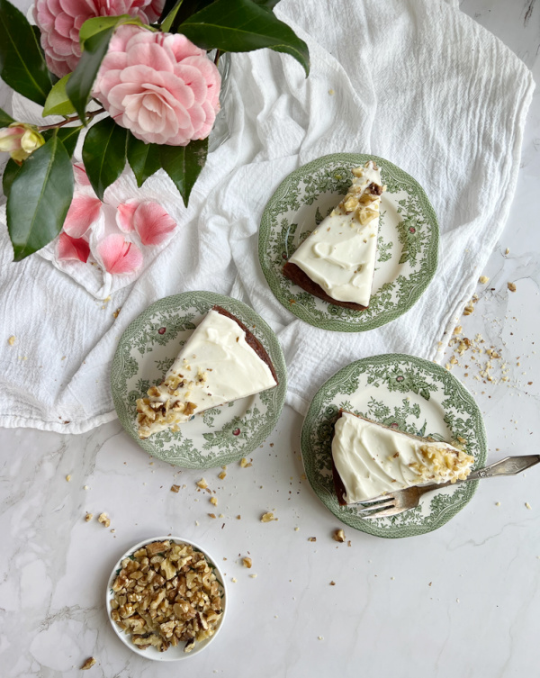 Ina Garten Old Fashioned Banana Cake with cream cheese frosting in 3 pieces on green and white transfer ware with a vase of camilla flowers. 