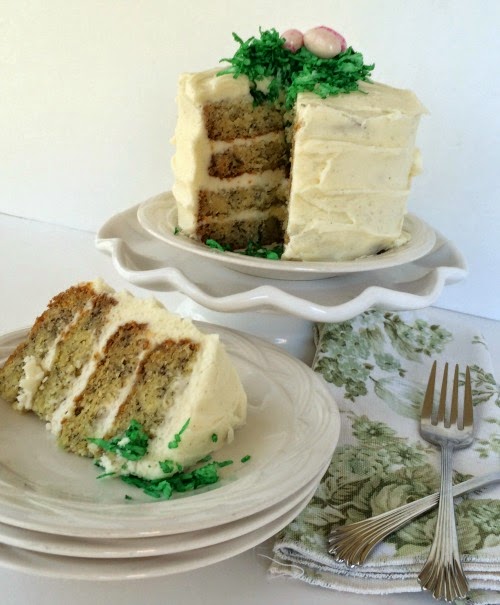 Banana cake frosted with cream cheese frosting and green coconut nut nest and 3 egg candies laying on a green and white toile napkin. 
