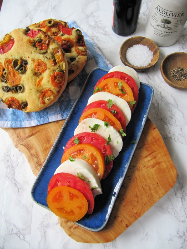 Herbed tomato and olive focaccia bread on a wood board with a side of tomato and mozzarella salad. 