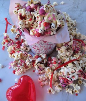 Bucket of chocolate covered popcorn with colored candy and sprinkles and a red plastic heart sitting next to it.