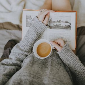 A woman reading in bed holding a cup of coffee.