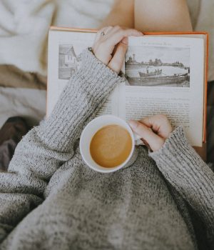 A woman reading in bed holding a cup of coffee.