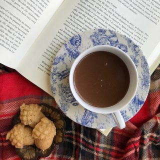 cup of hot chocolate in a blue and white toile cup and saucer laying on top of an opened book with a plaid throw and acorn cakelets