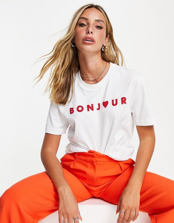 Giril with blog hair sitting on a white box wearing orange pants and a white t-shirt that has the words Bonjour on the front