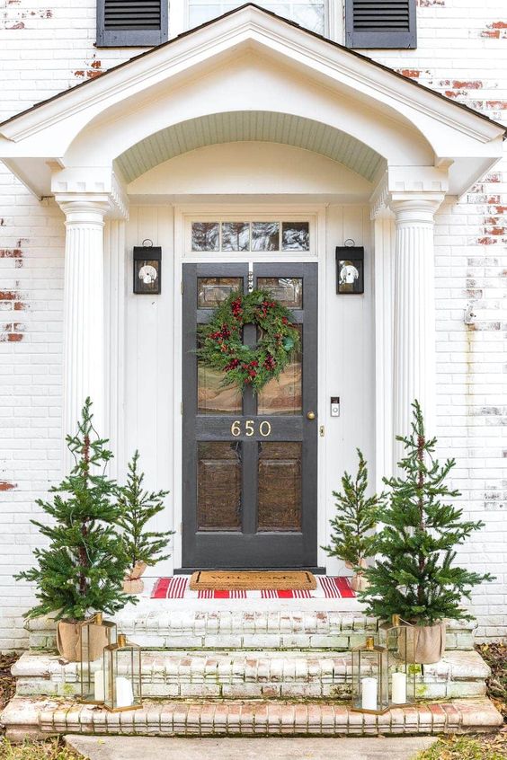 White distressed brick house with black door that has a green wreath and 4 little christmas trees