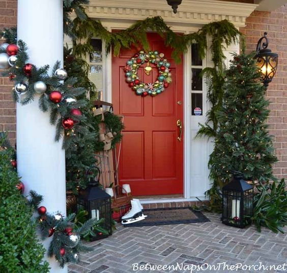 Red door with a wreath and a christmas tree on the right of the door, white columns wrapped in pine garland and a pair of ice skates to the left