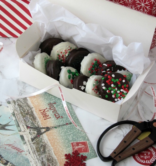 chocolate covered Oreos in a white box sitting on red and white stripped paper with postcards from Paris and a pair od scissors nearby. 