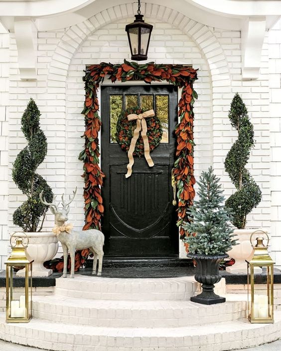 White brick house with magnolia leaf garland around the door and urn with a tree, two topiary in urns and two gold lanters