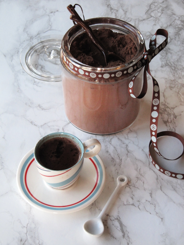 homemade-hot-chocolate-mix in a glass canister with a brown ribbon around the top and a striped cup filled with hot chocolate