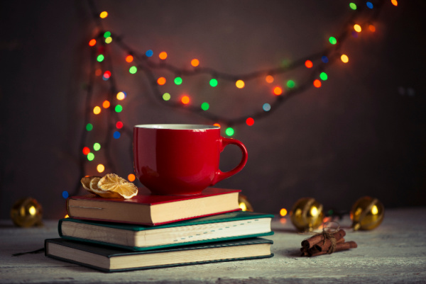 pile of books with a red cup of tea on top and colorful christmas light strung up behind them