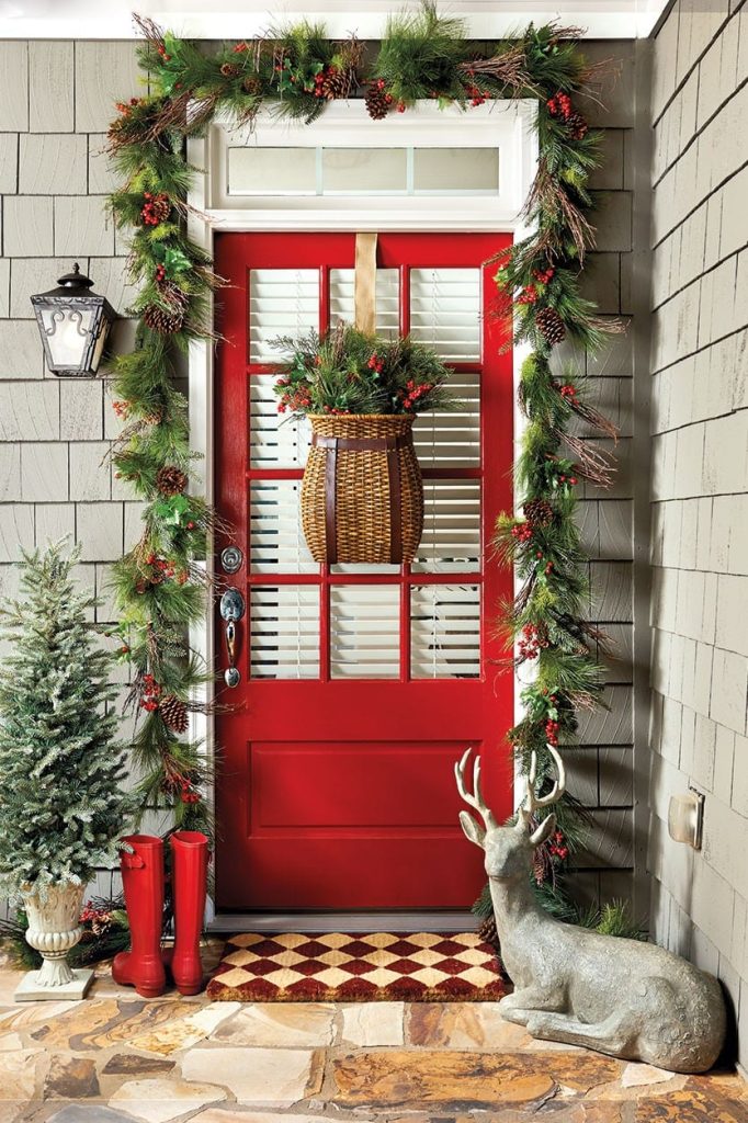 Red door with a wicket basket filled with greens. A garland around the door and a pair of small chrstmas trees on the leftt go the door with a pair of red boots and a small deer on the right side