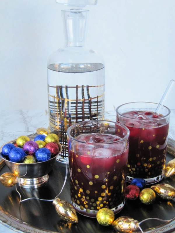 two cranberry cocktails in a clear glass with gold dots sitting on a silver tray with a decanter in the background and a small bowl with colored round chocolates