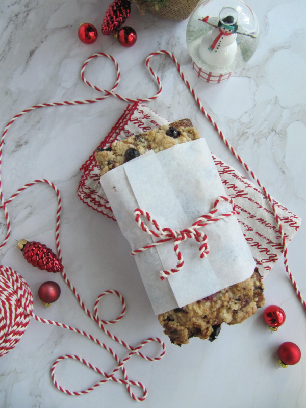 cranberry walnut loaf wrapped in white paper and tied with red and white string on a white piece of marble with scattered christmas bulbs a snow globe and string
