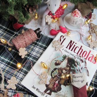 flatly of St.Nicholas books and a small bonus with a cup of hot chocolate on a green plaid background with gingerbread men and christmas lights.