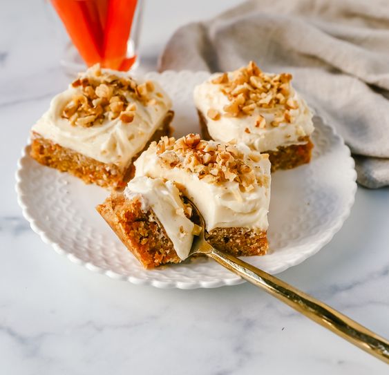 A white plate with three carrot cake bars one with a fork laying nearby in the background is a glass of carrots