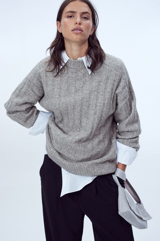 woman with dark hair wearing a pair of black pants, white shirt and an oversize gray sweater