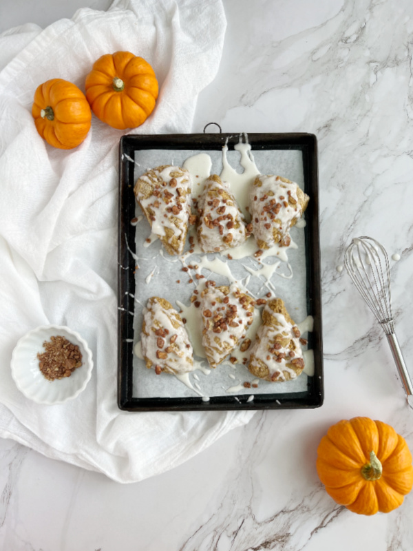 pumpkin scones on a black baking sheet with a whisk and pumpkins surrounding it