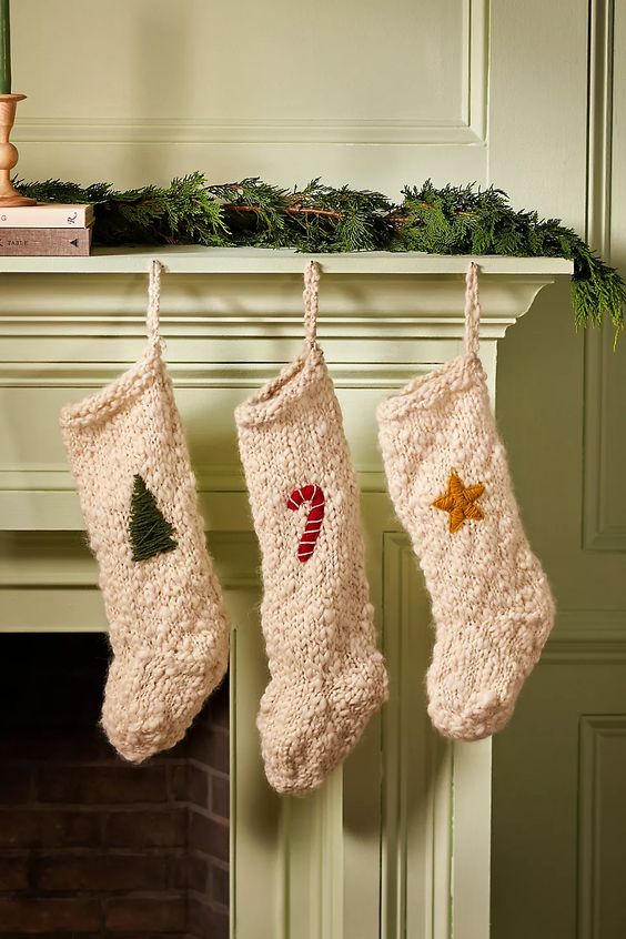 three white wool stockings, one with a green tree, one a candy cane and the other a star, hanging on a green mantle with a garland of greenery anthropologie.