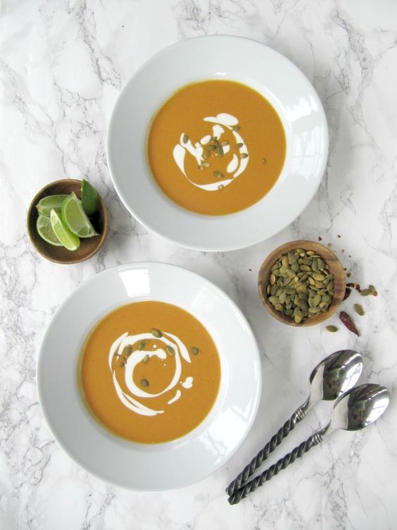 2 white bowls with pumpkins soup and a swirl of white cream alongside are two small bowls with limes and seeds and 2 spoons. 