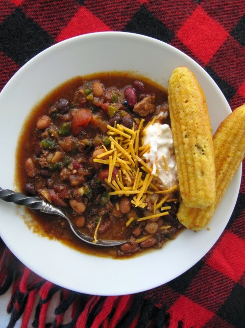 a white bowl of chili with two sticks of cornbread on the side of the bowl sitting on a black and red quilt. 