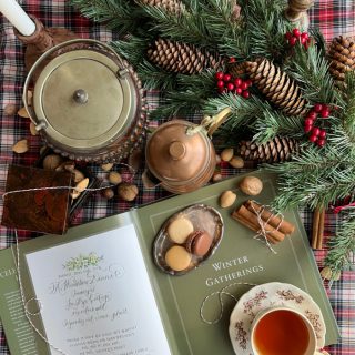 A flatlay photograph of a book opened to a page that says winter gatherings, with a cup of tea and scattered nuts and pinecones and evergreens.