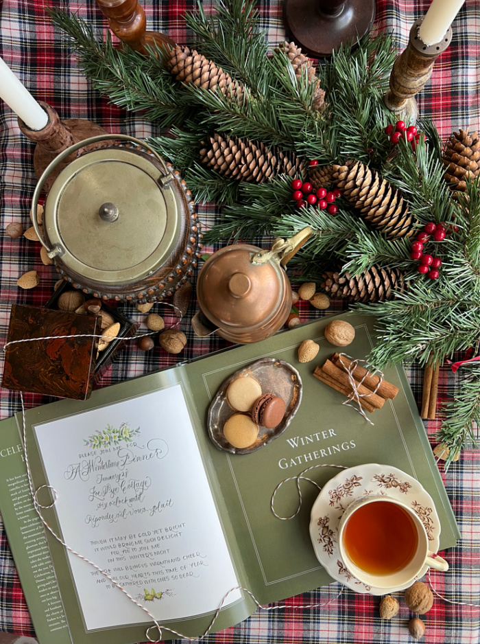 A flatlay photograph of a book opened to a page that says winter gatherings, with a cup of tea and scattered nuts and pinecones and evergreens. 