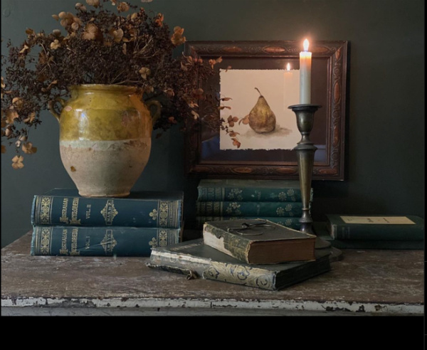 A hello and white pot on two books next to 3 books and a candle with a painting of a pear. 