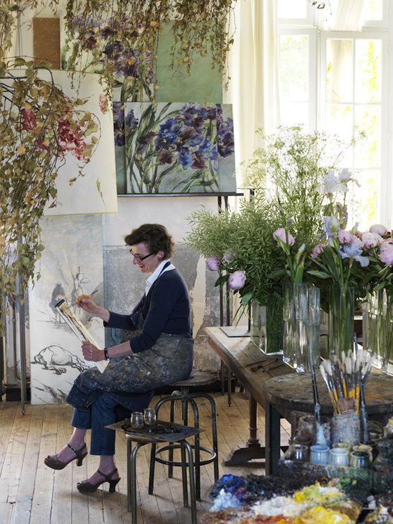 Artist Clair Basler sitting at her easel surrounded by flowers. 