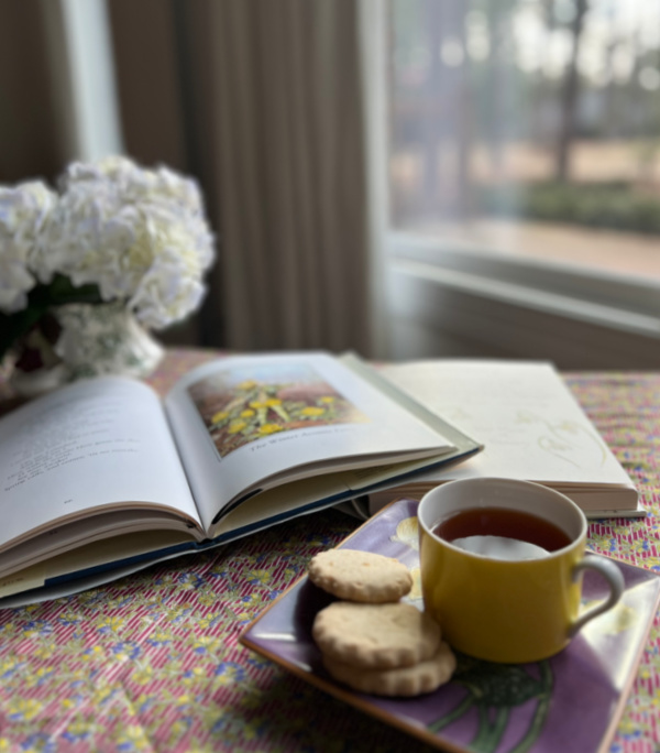 Cup of tea and books next to a window with a vase of flowers. 