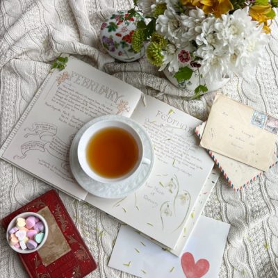 Flatlay photo with an open book a cup of tea, a vase of flowers and a small bowl of candy hearts alongside vintage letter.