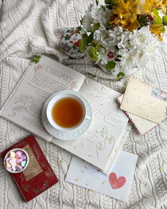 Flatlay photo with an open book a cup of tea, a vase of flowers and a small bowl of candy hearts alongside vintage letter. 