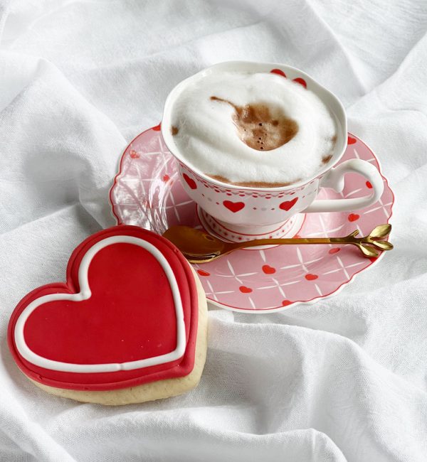 cup of hot chocolate with foam in a white cup with hearts on a pink saucer with a heart cookie.
