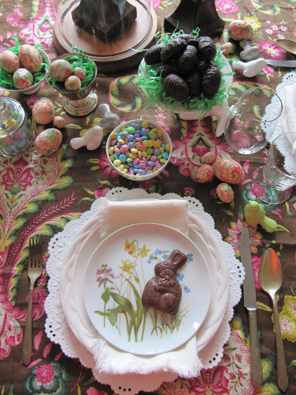 Easter table setting with white plates, pink and brown paisley tablecloth and lots of chocolate Easter eggs and bunnies. 