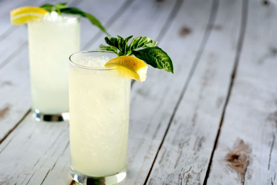 2 high ball glasses with a lemon cocktail and lemons and basil on the side. 