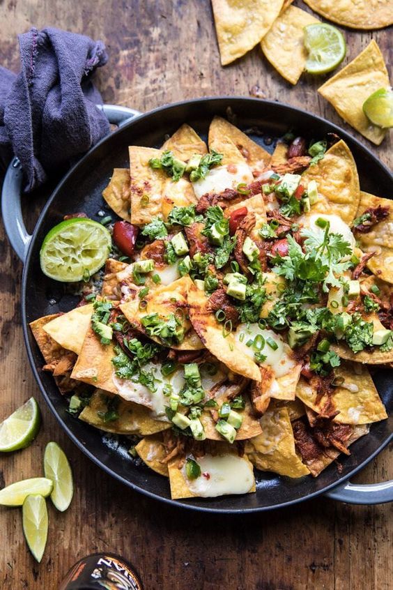 Chipotle Braised Chicken Nachos in a skillet with chips and limes scattered on the sides. 