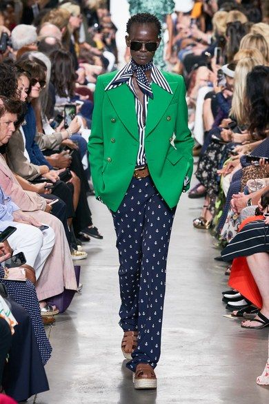 Woman walking the catwalk with a blue and white t-shirt and a blue and white pair of pants with a green blazer. 