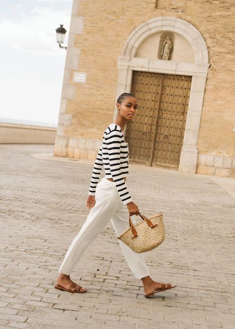 Woman in a blue and white shirt and white pants earring a wicker tote. 