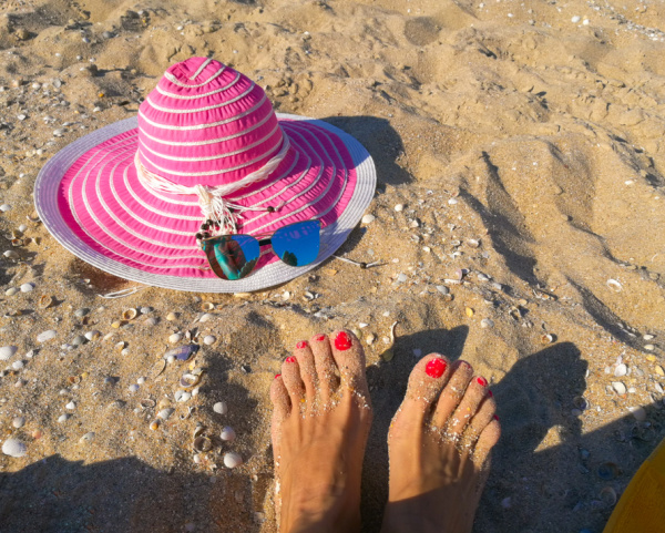 A pink sun hat laying in the sand near 2 feet with painted red toenails. 