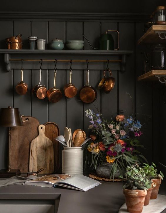 Dark green bead board wall with hanging copper pots above a counter with wood cutting boards and flowers. 