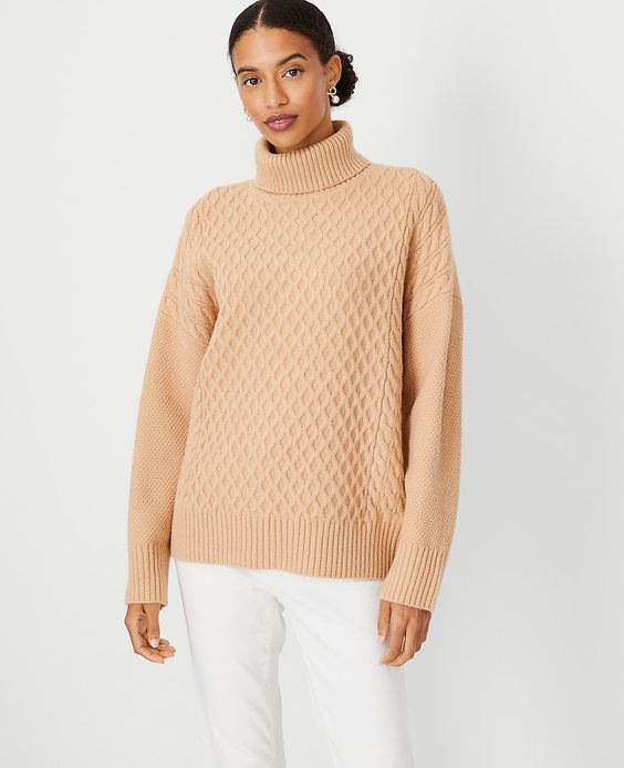 Woman wearing white pants and a camel colored cable knot sweater. 