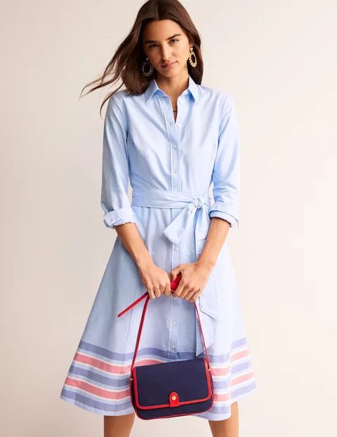 Woman wearing a pale blue shirt dress with red and blue stripe hem. 
