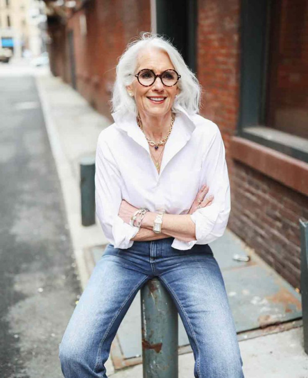 Woman in a white blouse and jeans with silver hair and glasses. 