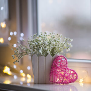 woven pink heart by a window with fairy lights and a white pot of flowers.