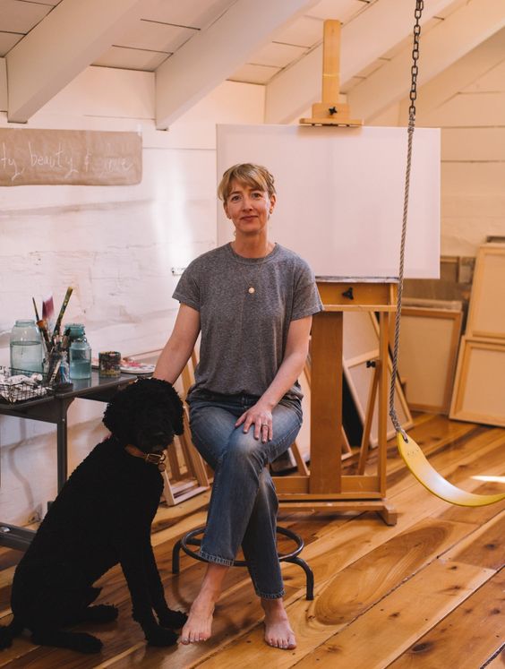 Kare Lewis Artist and Illustrator, sitting on a chair in front of an easel with a black dog. 