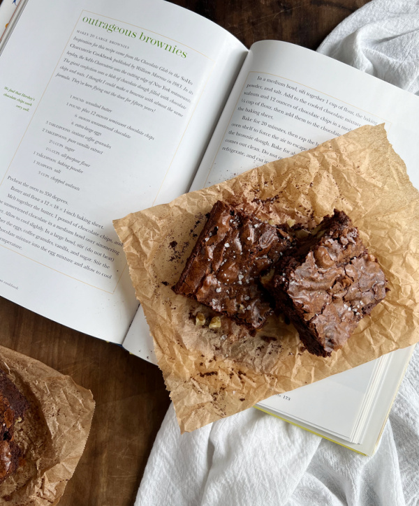 Two brownies on a brown piece of parchment sitting on an open cookbook with the recipe for outrageous brownies. 