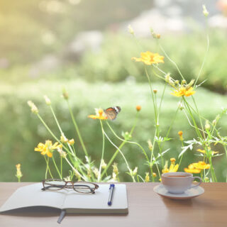 cup of tea in a white cup sitting next to a notebook with a pen and glasses with yellow flowers and a butterfly