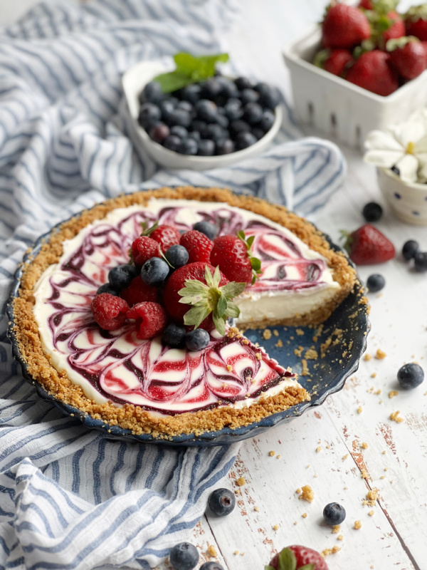 Red White and Blue No-Bake Cheesecake slices on a white plate with raspberries. in a white basket and blueberries in a white cup.