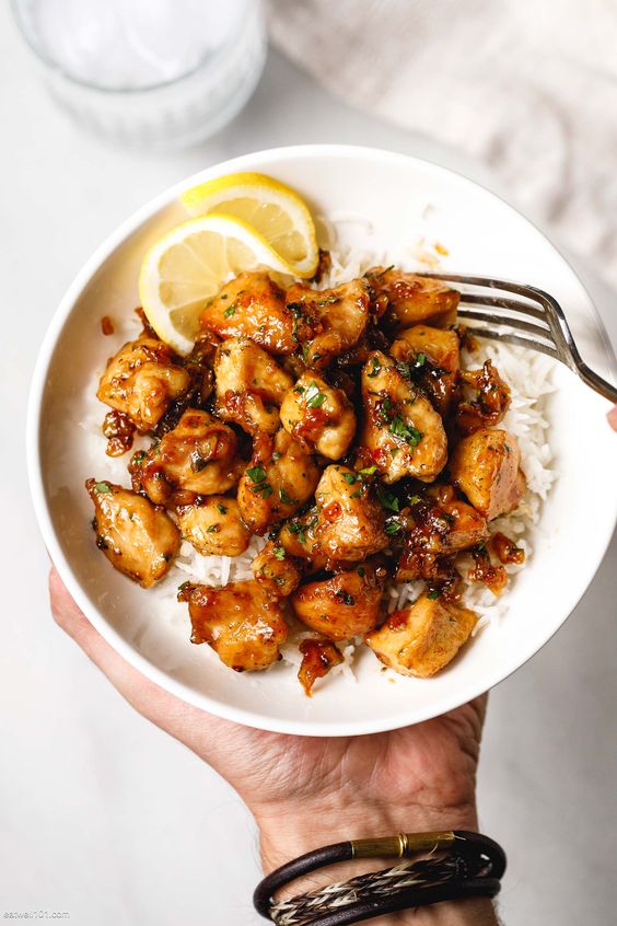 Honey garlic chicken bites over white rice in a white bowl with 2 slices of lime. 