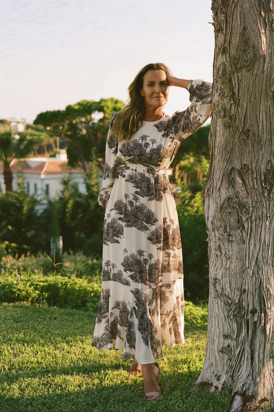 A woman standing next to a tree in a black and white toile dress. 
