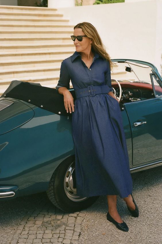 Woman in a blue shirt dress leaning on a vintage car. 